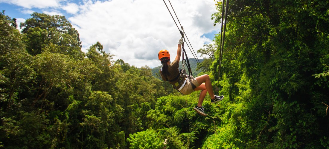 Person mit Helm und Sicherheitsgurt fährt an einer Zipline durch den tropischen Dschungel. Unten dichter grüner Wald, blauer Himmel mit Wolken im Hintergrund