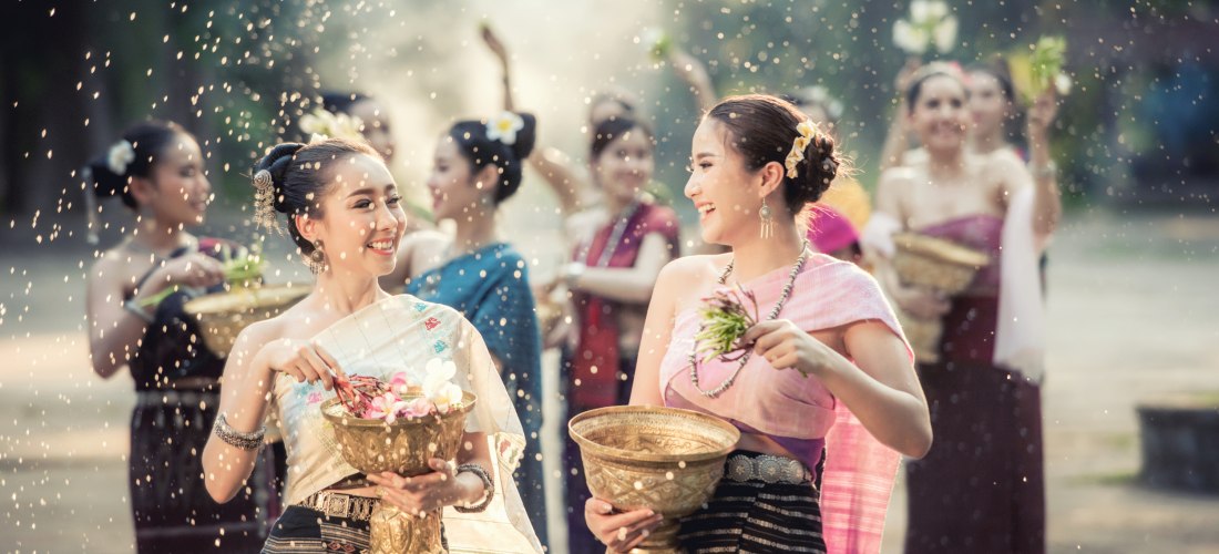Junge Frauen in traditioneller thailändischer Kleidung feiern mit Blumen und goldenen Schalen. Sie lächeln einander an, während Wasser in der Luft glitzert. Weitere Frauen im Hintergrund feiern mit.