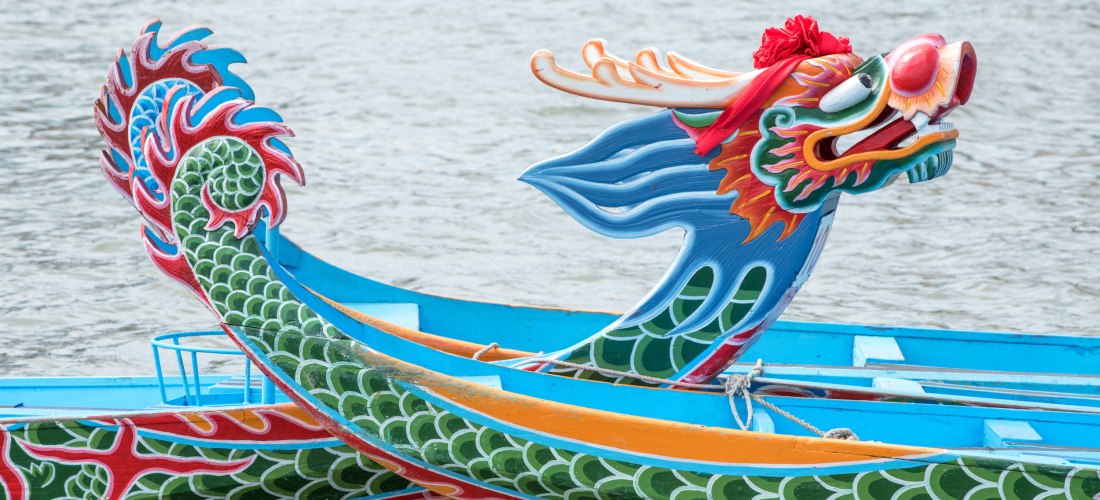Nahaufnahme eines Drachenboots mit leuchtend buntem Drachenkopf. Die Schnitzerei zeigt große Augen, Zähne und eine rote Zunge, geschmückt mit einer roten Blume. Das Boot liegt ruhig im Wasser.
