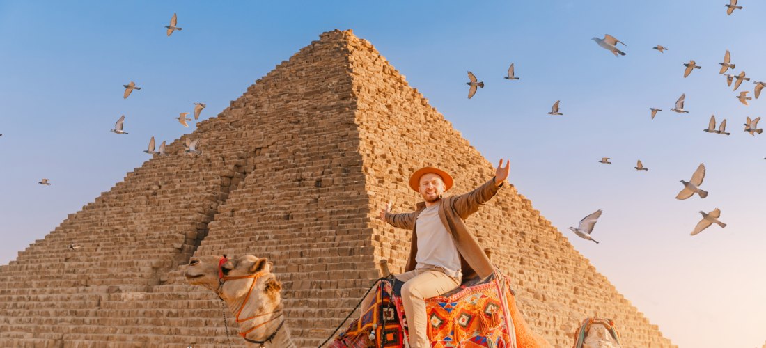 Ein lachender Tourist sitzt auf einem bunt geschmückten Kamel vor der großen Pyramide von Gizeh. Am Himmel fliegt ein Schwarm Tauben, der Himmel ist klar und blau