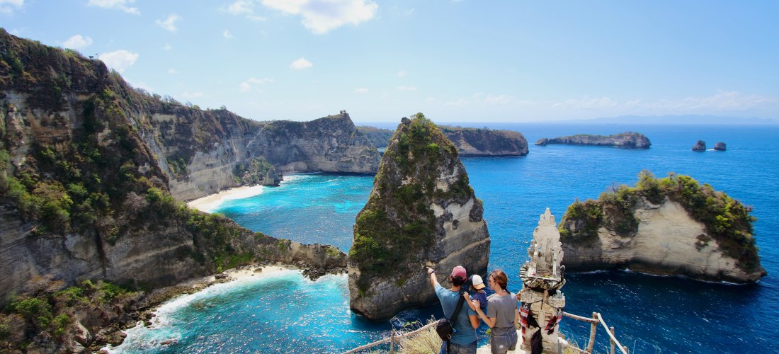 Eine Familie mit Kind steht an einem Aussichtspunkt hoch über dem Meer auf Nusa Penida, Bali. Der Blick fällt auf markante, steile Klippen und türkisblaues Wasser mit kleinen Stränden. Im Vordergrund befindet sich ein kleiner, reich verzierter balinesisch