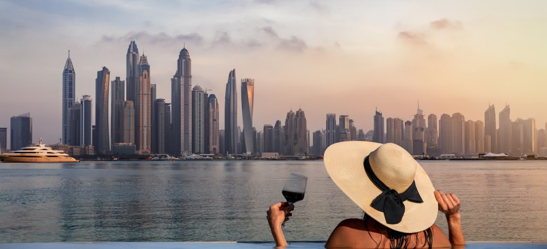 Eine Frau mit Hut und einem Glas Wein in der Hand steht am Rand eines Infinity Pools und schaut auf die Skyline der Dubai Marina