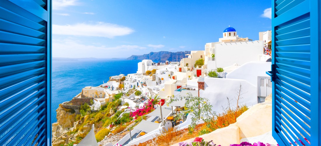 Blick durch ein offenes blaues Fenster auf die strahlend weißen Häuser von Santorini mit ihren typischen Kuppeln und bunten Türen. Im Vordergrund blühen leuchtend pinke, gelbe und weiße Blumen. Dahinter erstreckt sich das tiefblaue Meer bis zum Horizont u