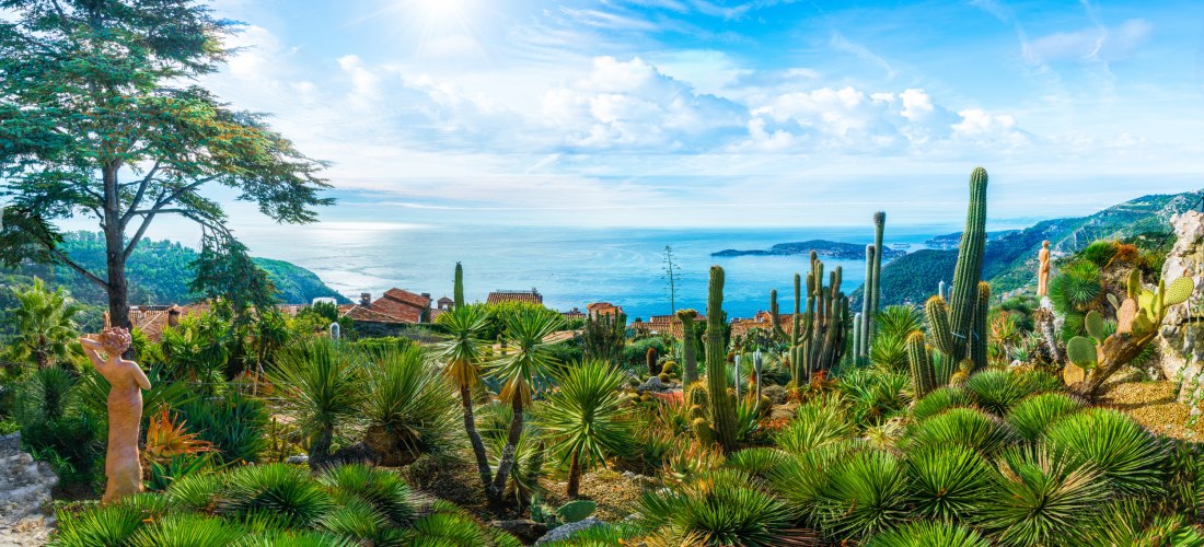 Mediterraner Garten mit Kakteen, Palmen und Skulpturen; im Hintergrund Blick auf die Küste und das blaue Mittelmeer.