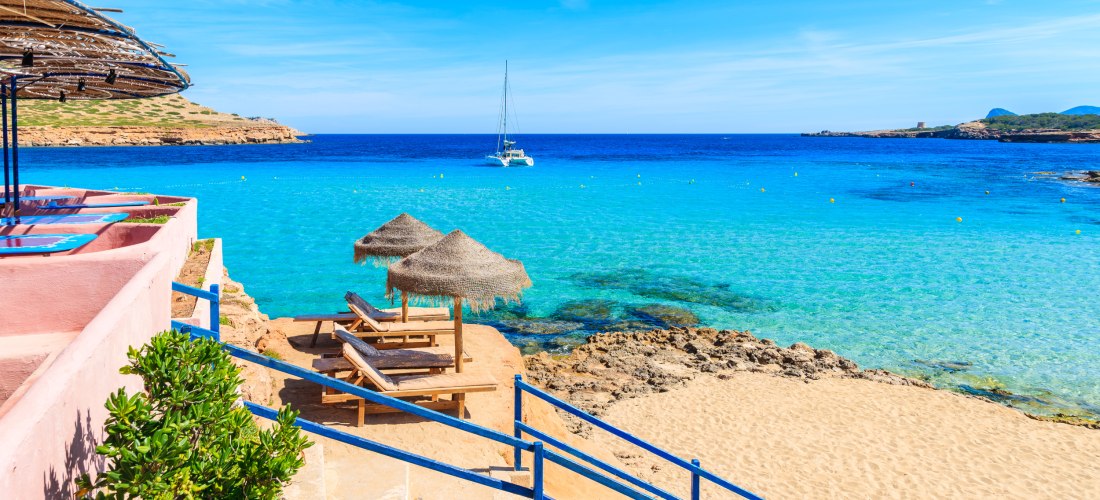 Blick auf den Strand Cala Comte auf Ibiza mit goldenem Sand, türkisblauem Wasser und zwei Sonnenliegen mit Strohdach. Im Hintergrund segelt ein weißes Boot auf dem tiefblauen Meer.