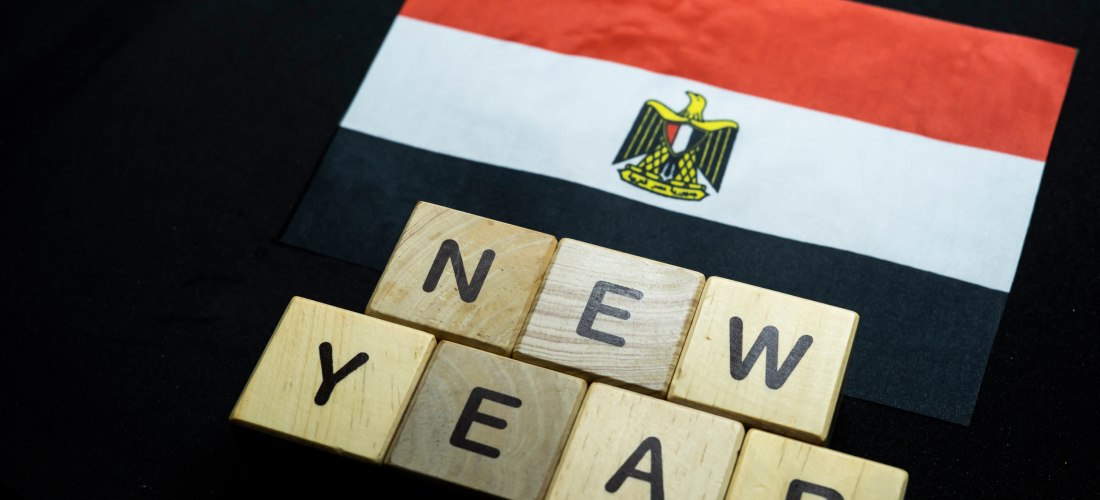 Ein rechteckiger Ausschnitt zeigt ein kleines ägyptisches Papierfähnchen auf schwarzem Hintergrund. Darunter liegen Holzbuchstaben, die das Wort 'NEW YEAR' bilden.