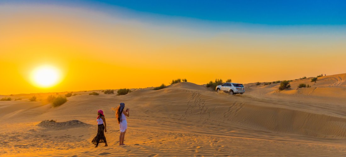 Zwei Frauen gehen bei Sonnenuntergang durch die Sanddünen einer Wüste, während ein Geländewagen im Hintergrund durch den Sand fährt. Der Himmel leuchtet in kräftigen Gelb- und Blautönen