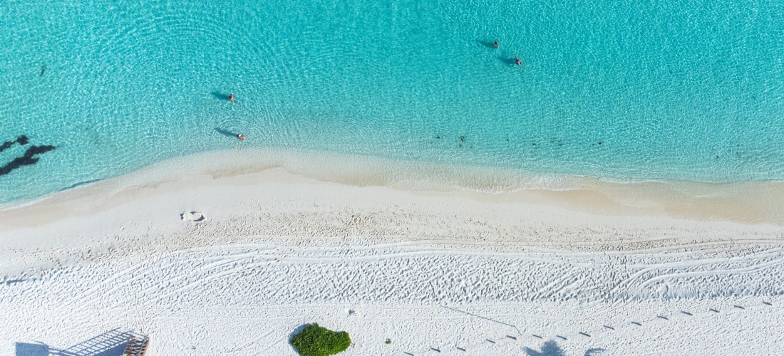 Ein traumhafter Sandstrand mit kristallklarem, türkisfarbenem Wasser aus der Vogelperspektive. Menschen genießen das ruhige Meer, während Palmen ihre Schatten auf den feinen weißen Sand werfen.