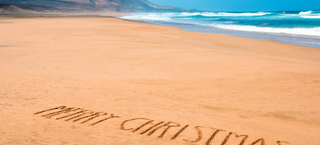 Weiter Sandstrand mit dem Schriftzug 'Merry Christmas' im Sand. Im Hintergrund Wellen und ein Bergmassiv unter blauem Himmel