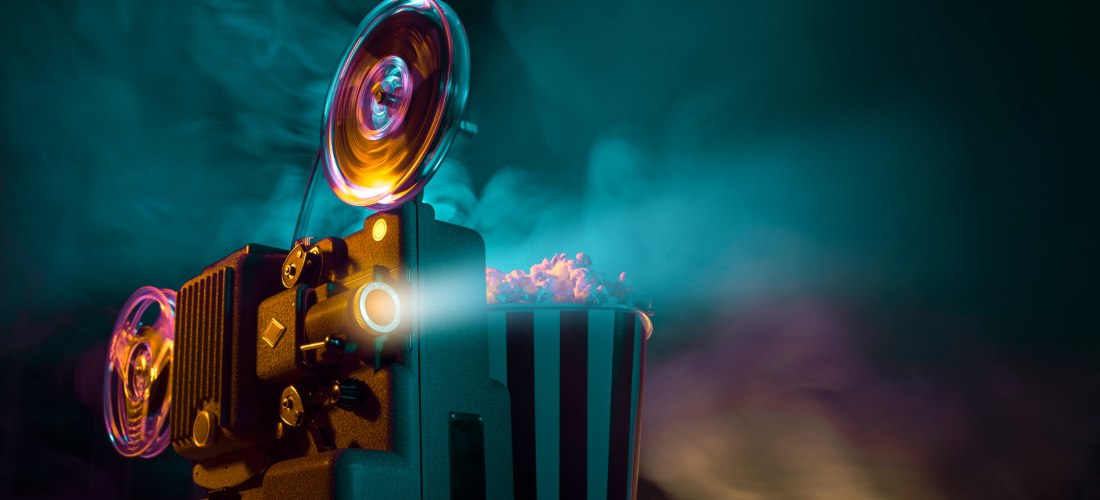 Ein alter Filmprojektor projiziert Lichtstrahlen durch den Raum. Daneben steht eine gestreifte Popcorntüte, aus der Popcorn herausquillt. Der Hintergrund ist stimmungsvoll in blauen und violetten Nebel gehüllt