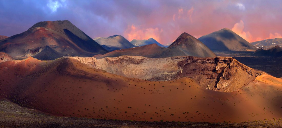 Weite Vulkanlandschaft mit rötlich-braunen Kratern und dunklem Lavagestein unter einem farbenprächtigen Himmel bei Sonnenuntergang. Im Hintergrund ragen weitere Vulkankegel auf.