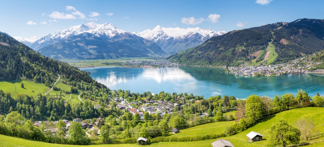 Weite Ansicht über grüne Almwiesen auf einen großen See in einem Tal; am Ufer eine Stadt, ringsum bewaldete Hänge und dahinter schneebedeckte Alpengipfel unter blauem Himmel.