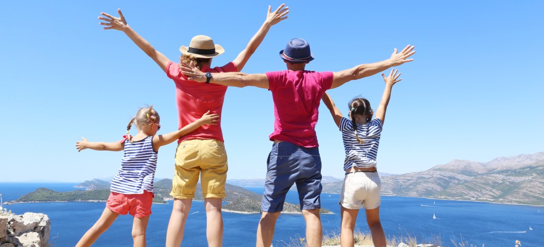 Eine Familie mit zwei Kindern steht auf einem Felsvorsprung mit Blick auf das Meer. Alle haben die Arme ausgebreitet und schauen fröhlich in die Ferne. Im Hintergrund sind das tiefblaue Meer, kleine Inseln und Berge zu sehen.