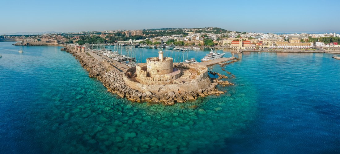 Der Mandraki-Hafen auf Rhodos: Im Vordergrund eine antike Säule mit einer Bronzestatue eines Hirsches, Symbol der Insel. Ein Angler sitzt am Rand des Kais. Im Hintergrund liegt die mittelalterliche Festung Agios Nikolaos, umgeben von Segelbooten und türki