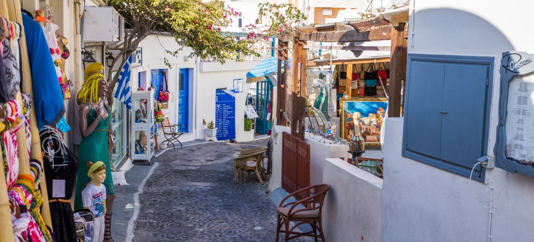 Eine enge, gepflasterte Gasse auf Santorini, gesäumt von Souvenirläden und kleinen Boutiquen. Links im Bild steht eine bunte Schaufensterpuppe mit gelber Perücke und grünem Kleid, daneben ein Kind mit Strohhut. Vor den Läden hängen bunte Kleider und Acces