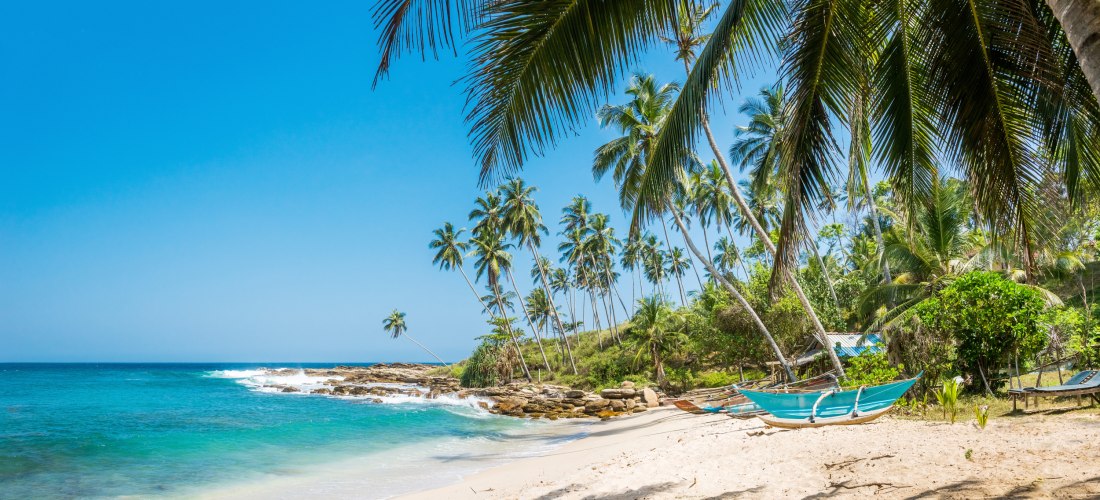 Panoramaaufnahme eines ruhigen tropischen Strandes mit weißem Sand, türkisblauem Meer und vielen Palmen. Im Vordergrund hängt ein traditionelles, blau gestrichenes Fischerboot zwischen zwei Palmen. Einige Wellen brechen leicht an den Felsen. Im Hintergrun