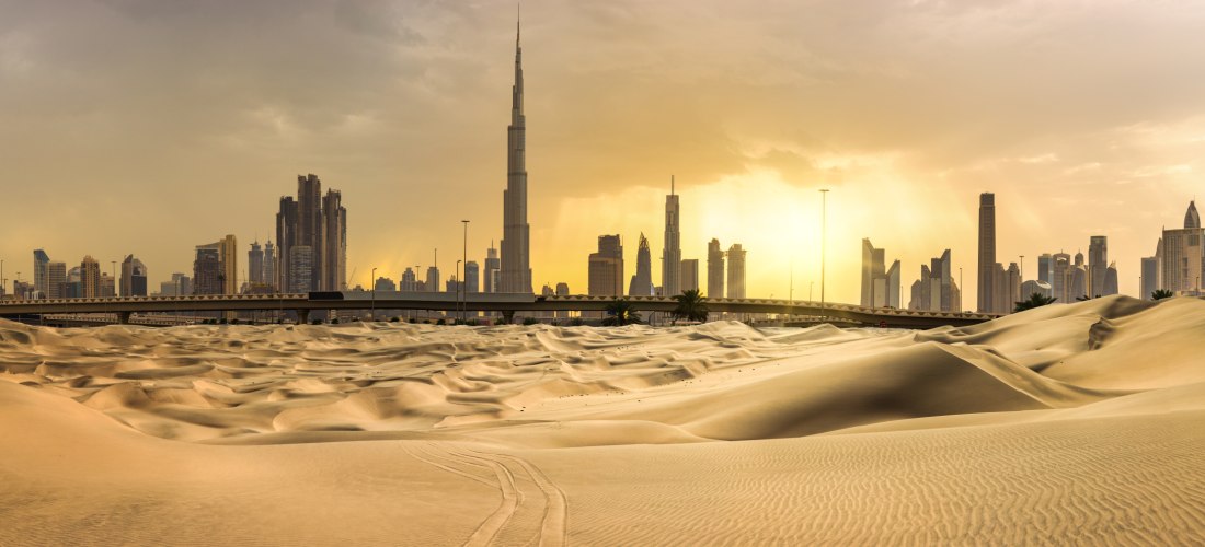 Dubai - Wüste Weite Wüstenlandschaft mit Sanddünen im Vordergrund und der Skyline von Dubai im Hintergrund, darunter das markante Burj Khalifa. Die Stadt wirkt durch den Sonnenuntergang in goldenes Licht getaucht. Die Kontraste zwischen Natur und moderner Architektur s