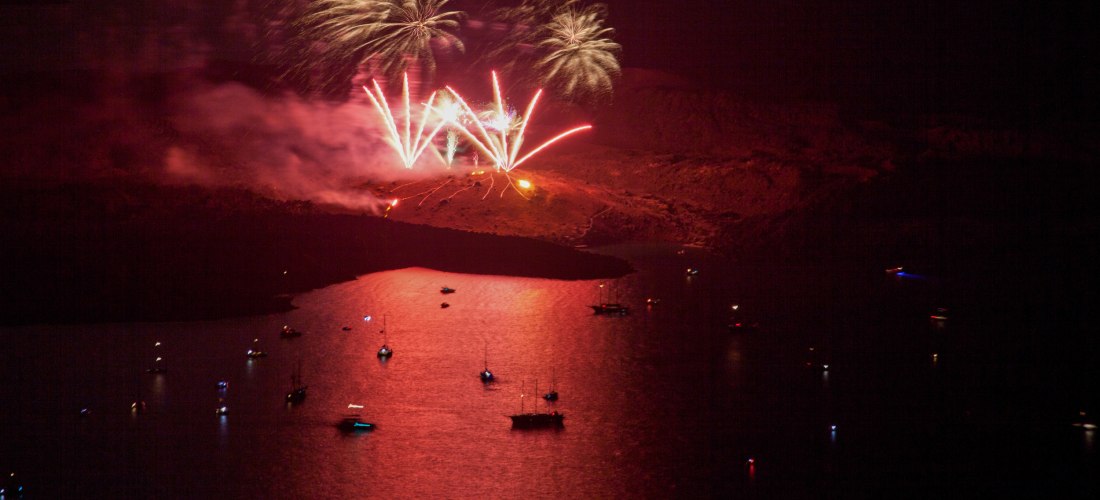 Nächtliche Szene auf Santorini mit einem spektakulären Feuerwerk über der Vulkaninsel Nea Kameni. Vom dunklen Himmel leuchten rote und goldene Raketen, während sich das Licht auf dem ruhigen Wasser der Caldera spiegelt. Mehrere kleine Boote beobachten das