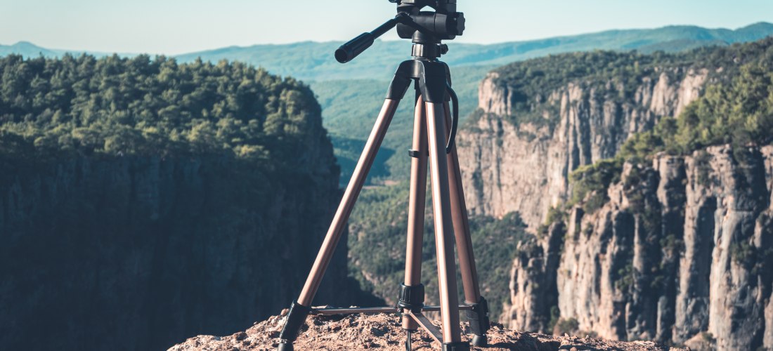 Ein Kamerastativ steht auf einem felsigen Aussichtspunkt mit Blick auf eine tiefe Schlucht und bewaldete Berge im Hintergrund.