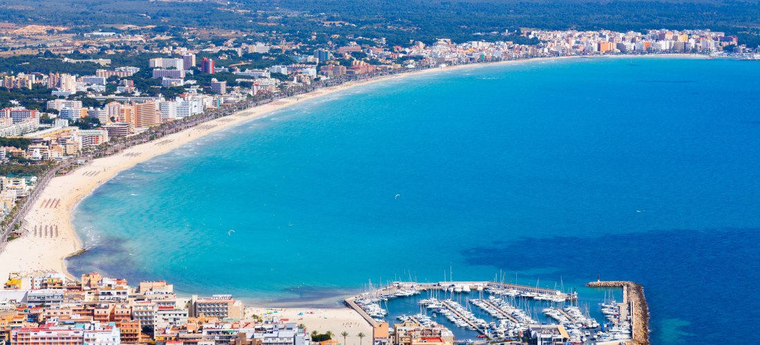 Luftaufnahme von Can Picafort im Norden Mallorcas. Entlang der Küste erstreckt sich ein heller Sandstrand, gesäumt von Hotels und Apartmentanlagen. Der Strand ist gut besucht, mit Sonnenschirmen und Liegen. Das Wasser ist türkisblau und klar, einige Boote