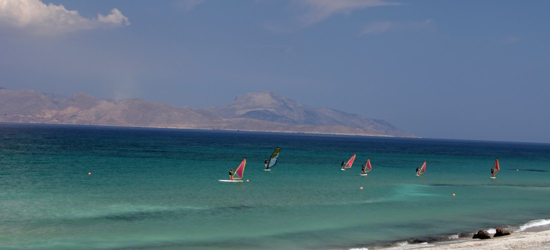 Mehrere Windsurfer gleiten auf türkisblauem Wasser vor der Küste von Kos. Der helle Sandstrand liegt ruhig im Vordergrund, dahinter ragen die Hügel und Berge einer benachbarten Insel am Horizont auf. Der Himmel ist klar mit wenigen Wolken