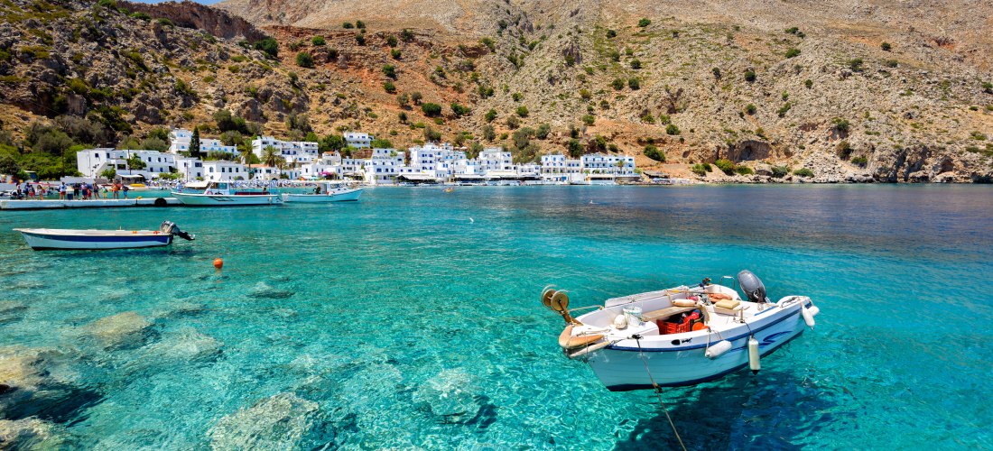 Klares türkisblaues Meer mit Booten vor einem kleinen, weißen Küstendorf am Fuße eines felsigen Berges auf Kreta