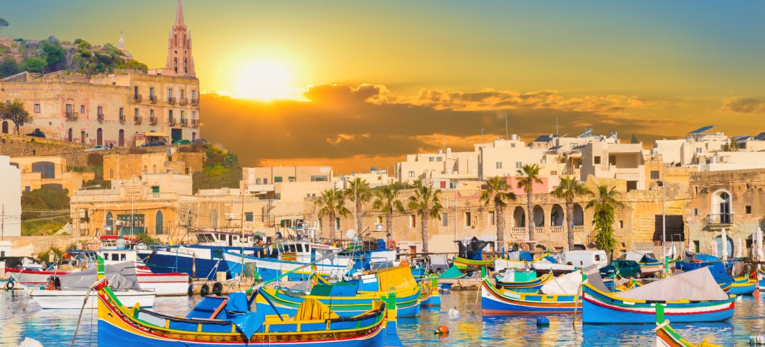 Bunte traditionelle Fischerboote (Luzzus) im Hafen von Marsaxlokk auf Malta bei Sonnenuntergang mit goldener Abendstimmung