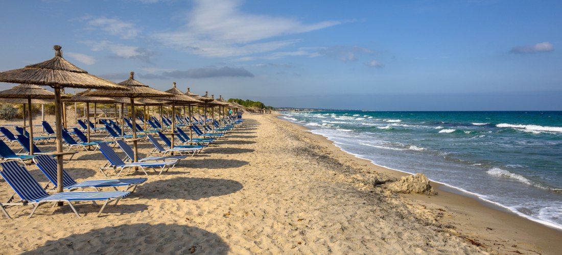 Das Bild zeigt einen langen, sandigen Strand mit zwei Reihen leerer Liegestühle. Die Liegen sind blau gestreift und stehen unter großen Sonnenschirmen mit Strohdach. Die runden Schatten der Schirme liegen auf dem Sand. Rechts im Bild trifft das türkisfarb
