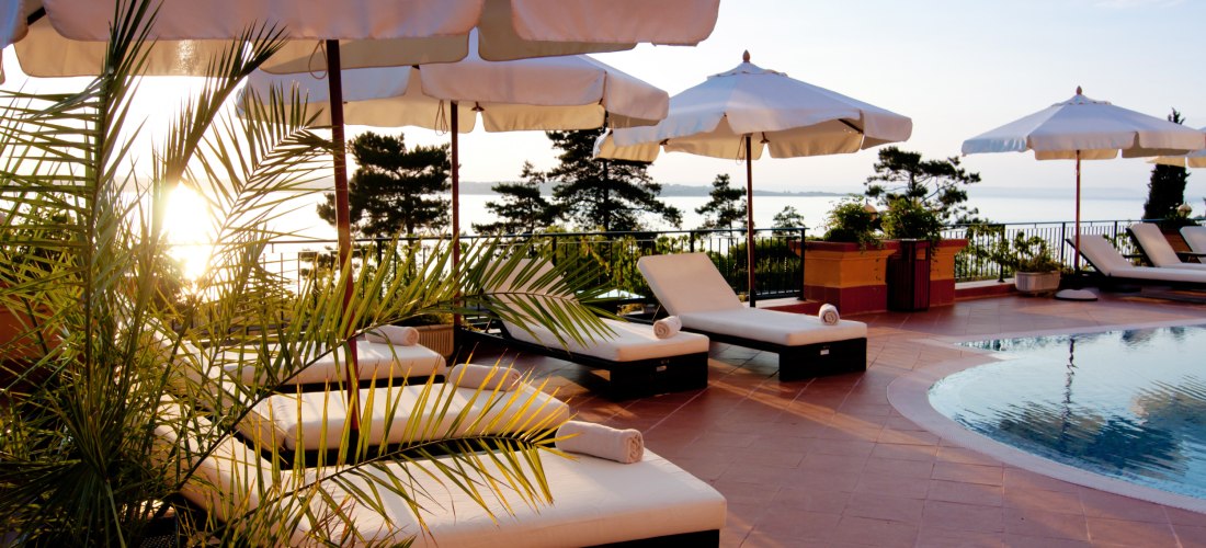 Sonnige Poolterrasse eines Resorts mit bequemen weißen Liegen, weißen Sonnenschirmen und Blick auf das glitzernde Meer im Hintergrund. Palmen säumen den Bereich.