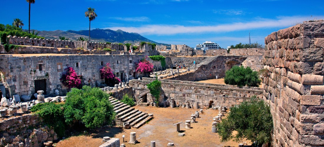 Mittelalterliche Festungsruinen auf Kos: massive Steinmauern, Stufen und verstreute Säulenfragmente, mit Bougainvillea und Palmen.