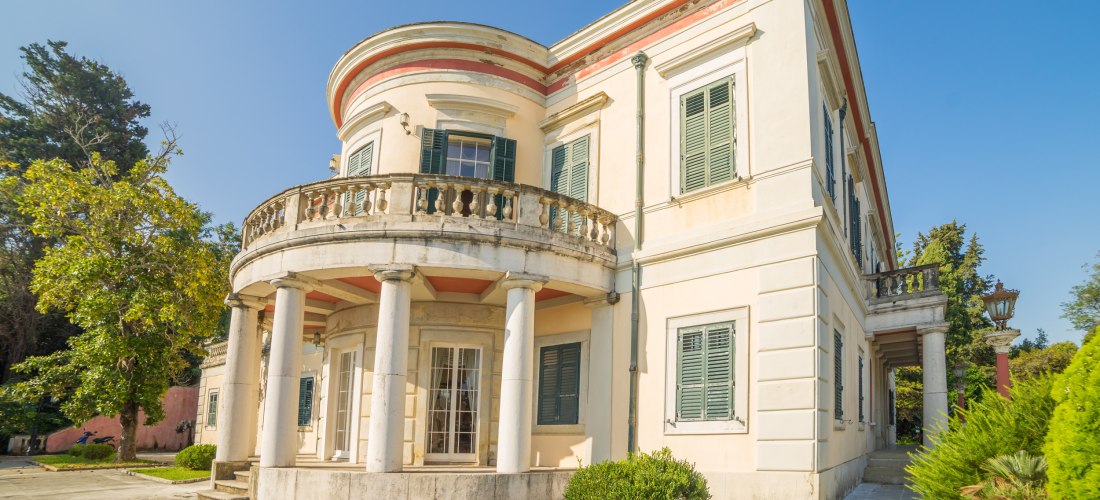 Eine elegante weiße Villa mit grünen Fensterläden, Rundbalkon und hohen Säulen im Eingangsbereich. Die Villa steht inmitten eines gepflegten Gartens mit Sträuchern und Bäumen, der Himmel ist klar und blau