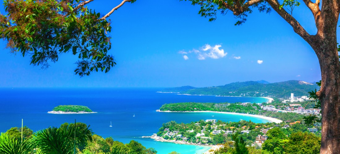 Panoramablick über die Südwestküste der thailändischen Insel Phuket. Im Vordergrund rahmen dichte grüne Bäume das Bild ein, mit einem großen Ast im oberen Teil. Unten sieht man mehrere türkisfarbene Buchten mit weißen Sandstränden und einer vorgelagerten 