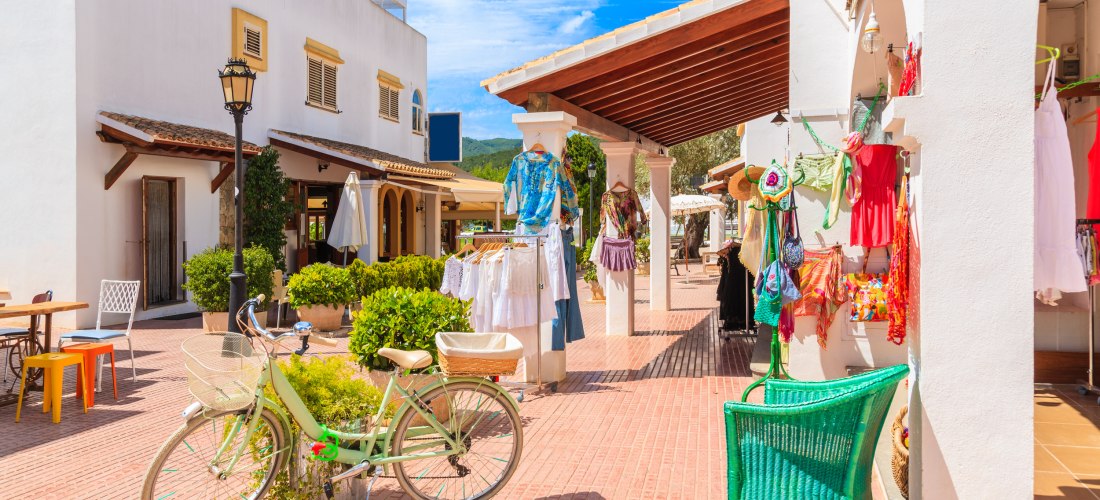 Blick auf eine sonnige Marktstraße in Ibiza mit weiß getünchten Häusern, bunter Kleidung an Ständern, einem grünen Stuhl und einem Fahrrad im Vordergrund.