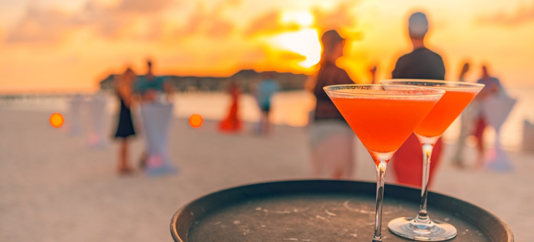 Zwei elegante Cocktailgläser auf einem Tablett mit verschwommener Strandparty und Sonnenuntergang im Hintergrund