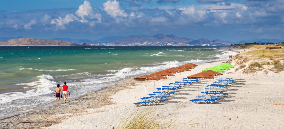 Zwei Personen spazieren am feinsandigen Strand entlang, das Meer brandet leicht ans Ufer. Rechts stehen Reihen von blauen Liegestühlen mit Sonnenschirmen, teils braun, teils grün. Im Hintergrund sind Hügel und Küstenlandschaft zu sehen, darüber ein dramat