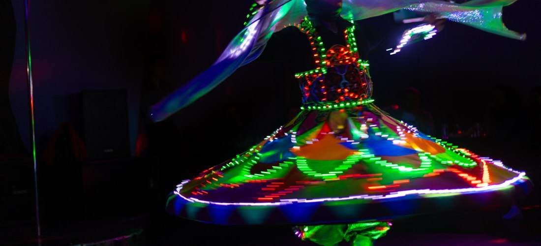 Ein Tanoura-Tänzer führt einen traditionellen ägyptischen Derwisch-Tanz auf. Er trägt ein farbenfrohes, weit ausgestelltes Kostüm, das mit leuchtenden LED-Lichtern geschmückt ist. Die Lichter erzeugen während der Drehung kreisförmige Muster. Die Szene ist