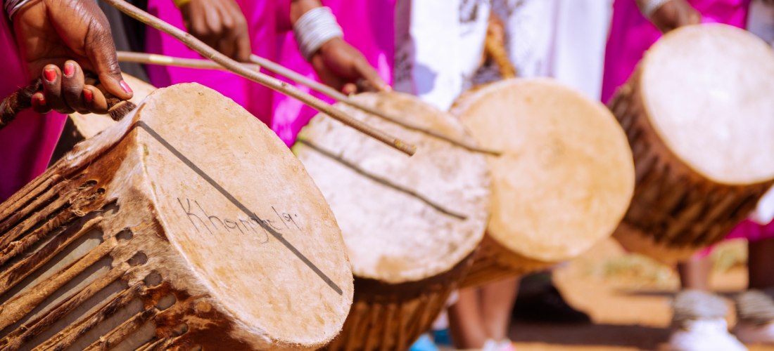 Mehrere Menschen schlagen mit Stöcken auf traditionelle afrikanische Trommeln. Die Hände sind mit bunten Armreifen geschmückt, im Hintergrund leuchten pinkfarbene Stoffe.