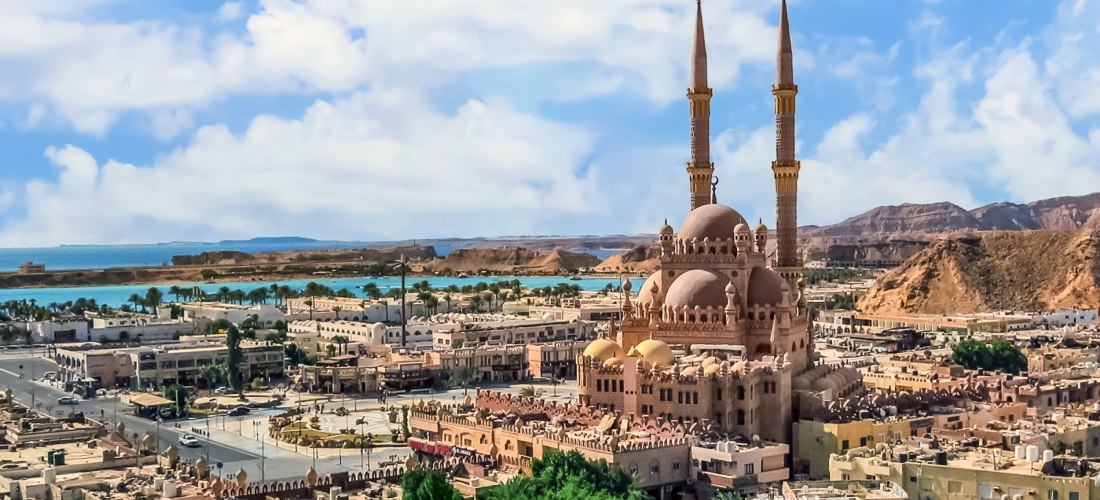 Panoramablick über die Stadt Sharm el Sheikh in Ägypten mit der imposanten Al Sahaba Moschee im Zentrum. Die Moschee hat zwei hohe Minarette und mehrere goldene Kuppeln. Sie ist von flachen Gebäuden, Straßen und grünen Palmen umgeben. Im Hintergrund sieht