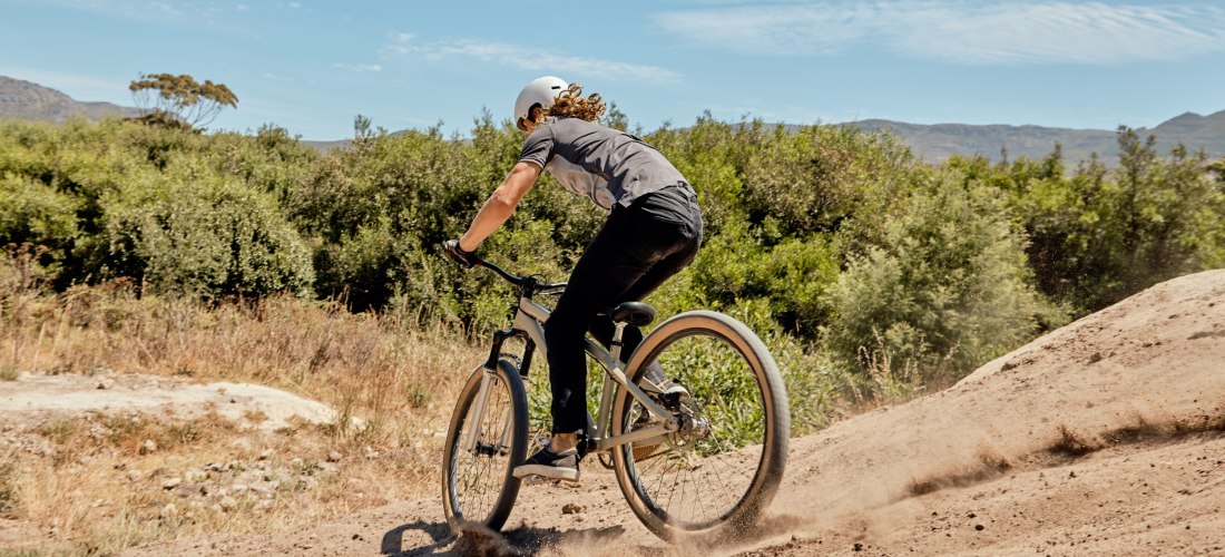 Eine Person fährt mit einem Mountainbike über eine staubige Piste, umgeben von grüner Buschlandschaft und unter einem leicht bewölkten Himmel. Die Szene vermittelt sportliche Aktivität und Naturverbundenheit.
