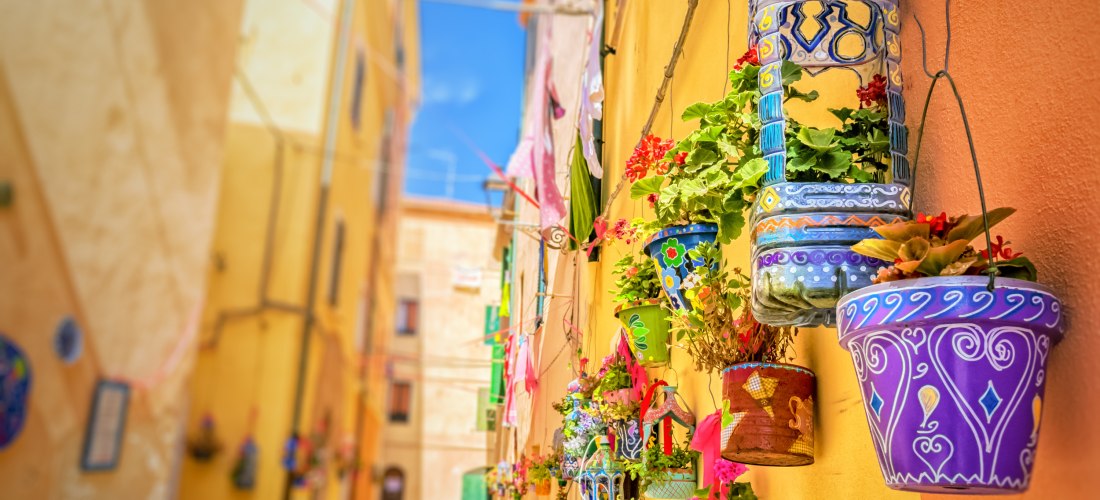 Farbenfrohe Gasse in der Altstadt von Alghero auf Sardinien mit bemalten Blumentöpfen und mediterraner Atmosphäre – beliebtes Reiseziel an der Westküste