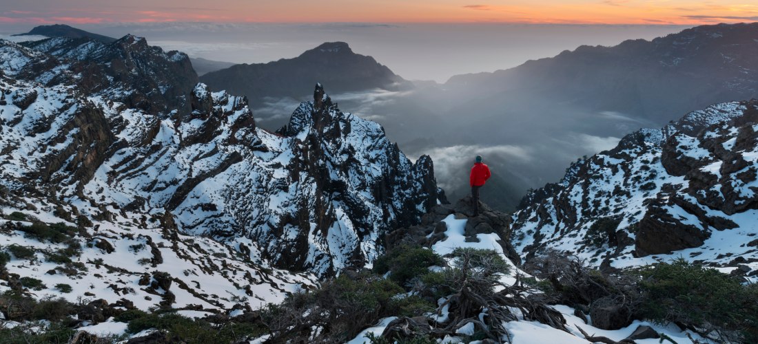 Person in roter Jacke steht auf einem verschneiten Bergkamm mit Blick über ein beeindruckendes Gebirgspanorama bei Sonnenuntergang. Die schroffen Felsen sind von Schnee bedeckt, Wolken hängen in den Tälern. Die Szene strahlt Ruhe, Weite und Naturerlebnis 