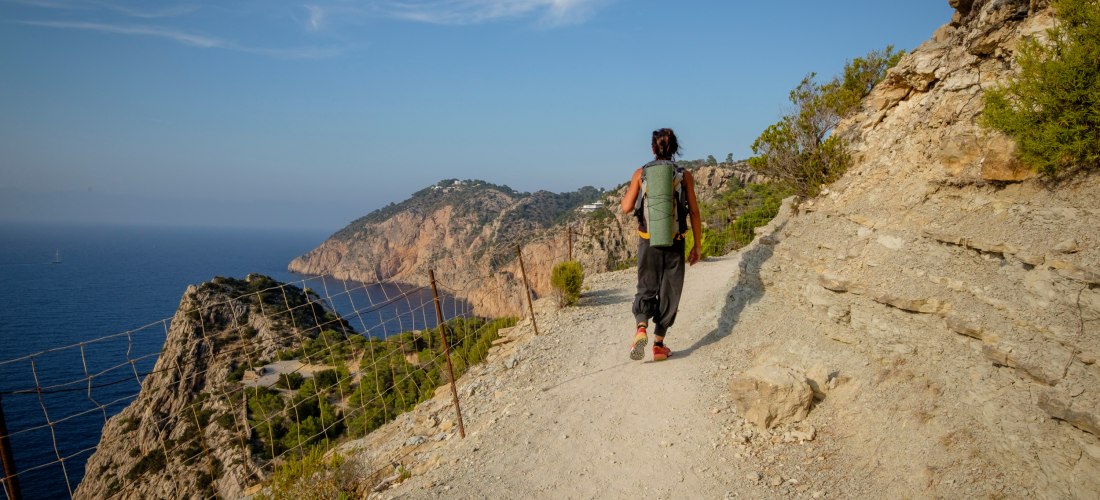 Eine Person mit Rucksack wandert auf einem schmalen, steinigen Küstenpfad entlang einer steilen Klippe. Rechts türmen sich Felswände, links fällt der Blick weit über das tiefblaue Mittelmeer und grüne Küstenlandschaften. Der Himmel ist blau mit ein paar S