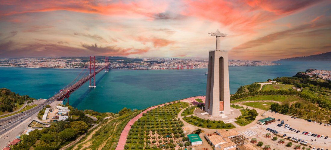 Luftaufnahme der Christusstatue Cristo Rei in Lissabon mit ausgebreiteten Armen, die über dem Fluss Tejo thront. Links führt die rote Hängebrücke Ponte 25 de Abril über das türkisfarbene Wasser in die Stadt. Im Hintergrund erstreckt sich die Skyline von L