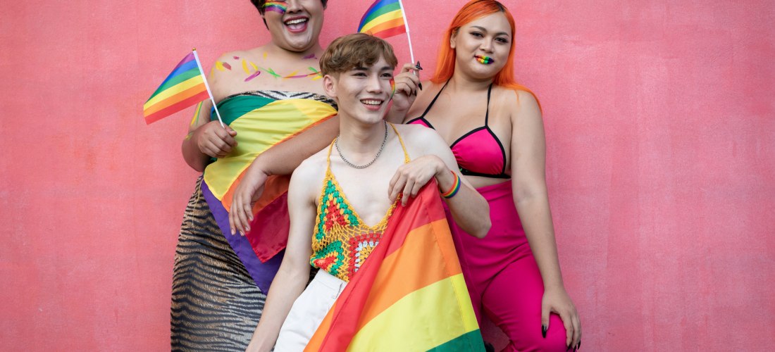 Das Bild zeigt drei Personen, die selbstbewusst und fröhlich vor einer pinkfarbenen Wand posieren. Sie tragen farbenfrohe Kleidung in Pride-Farben, darunter ein Regenbogenhäkel-Top, ein pinkes Outfit und ein Wickeltuch mit Regenbogenflagge. Zwei der Perso