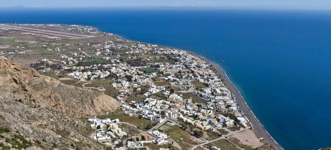 Kamari auf Santorini vom Mesa Vouno aus: Küstenort mit langem schwarzem Lavastrand, Häusern am Fuß des Berges und weitem Blick über die Ägäis