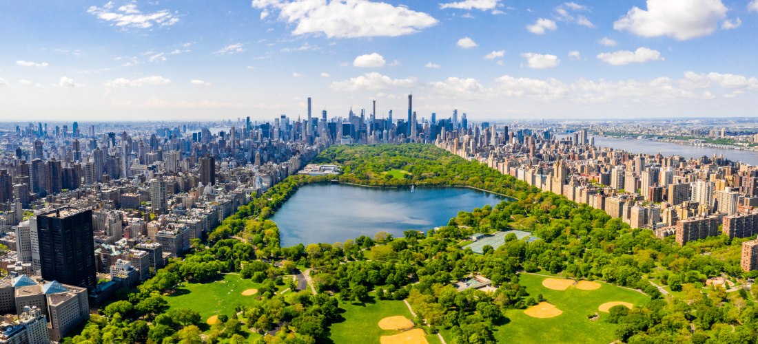 Luftaufnahme von New York City mit dem weitläufigen Central Park im Zentrum. Der Park ist von Hochhäusern und dicht bebauten Straßen eingerahmt. In der Ferne ist die Skyline von Manhattan mit zahlreichen Wolkenkratzern unter einem teils bewölkten blauen H