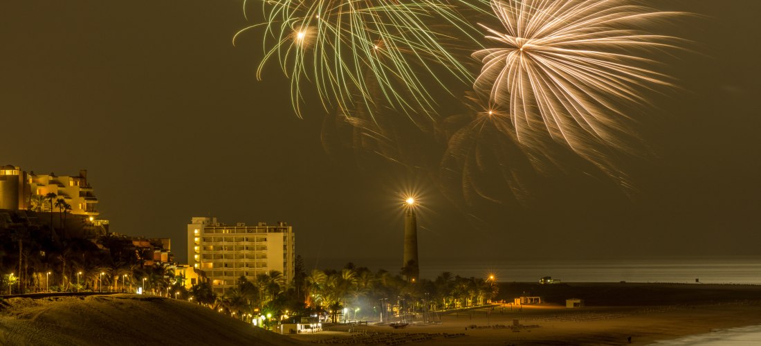 Feuerwerk über einem nächtlich beleuchteten Strand mit Leuchtturm und Hotels im Hintergrund.