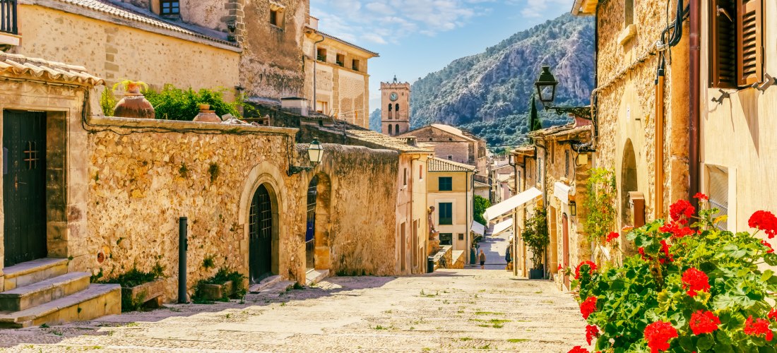Historische Altstadtgasse von Pollença auf Mallorca. Die Calvari-Treppe zieht sich mit Kopfsteinpflasterstufen bergauf durch die Altstadt, flankiert von Natursteinhäusern mit Holztüren und Fensterläden. Links und rechts blühen rote Geranien in Töpfen. Im 