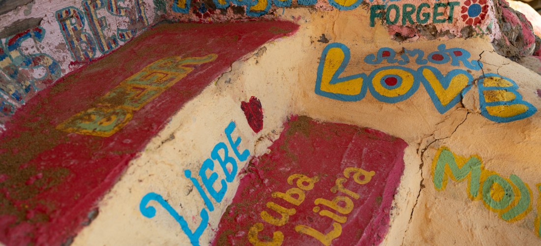Eine bemalte Steintreppe mit bunten Worten und Schriftzügen wie 'LOVE', 'MOJITO', 'Cuba Libra', 'LIEBE' und weiteren in verschiedenen Sprachen. Die Farben sind kräftig und lebendig – Rot, Gelb, Blau – und vermitteln ein künstlerisches, freies Flair. Die O