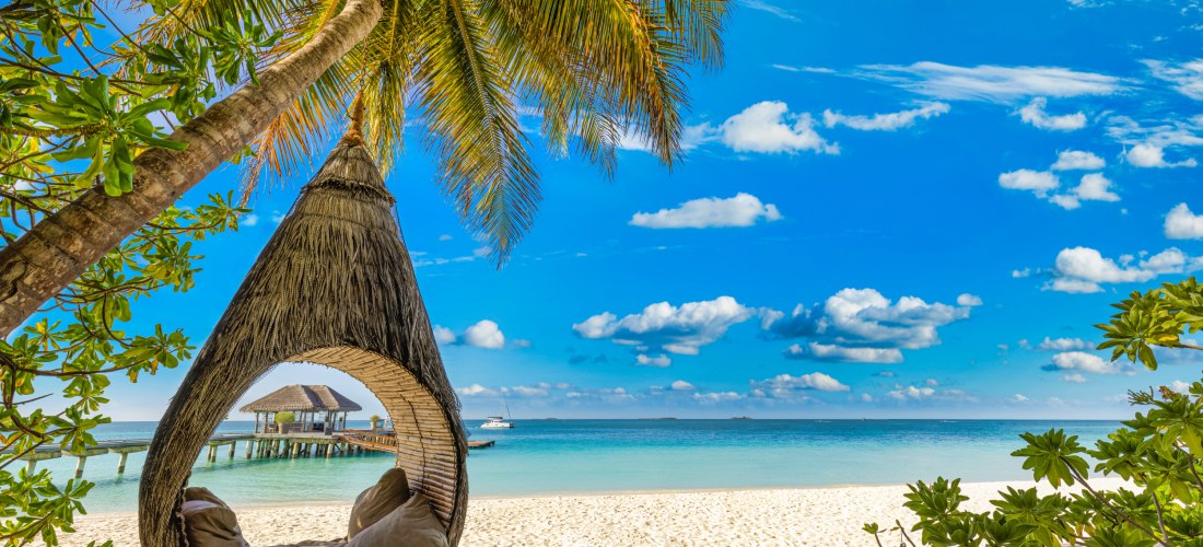 Hängender Kokon-Sessel an einer Palme mit Blick auf weißen Sandstrand, türkisblaues Meer und Pavillon auf dem Wasser bei sonnigem Himmel
